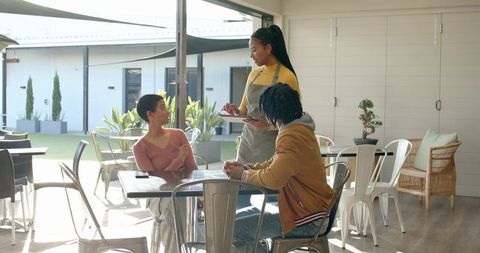 Female waitress taking order while couple chatting at sunlit courtyard cafe hospitality