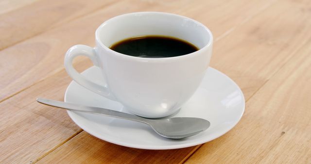 White ceramic cup filled with black coffee placed on a matching saucer. A silver spoon lies beside it on a light wooden table. Suitable for use in articles or advertisements related to coffee, breakfast scenes, cafes, or simple lifestyle settings.
