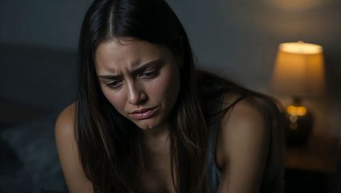 Young woman sitting on bed at night furrowing brows, bedside lamp glowing warmly
