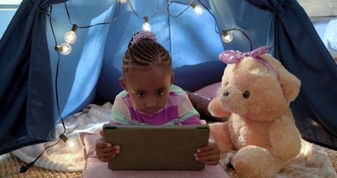 African american girl playing on tablet in cozy blanket fort with teddy bear and string lights