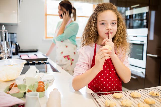 Young girl taking freshly baked cookies while her mother is distracted talking on her mobile phone in a modern home kitchen. Ideal for advertisements, articles about parenting, family life, home activities, baking, and playful moments.
