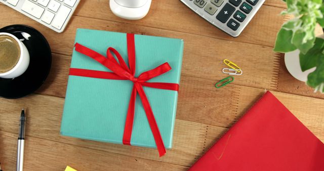 A neatly wrapped gift with a red ribbon sits on a wooden desk, surrounded by office supplies, with copy space. It suggests a festive or celebratory occasion in a workplace setting, such as a colleague's birthday or a work anniversary.