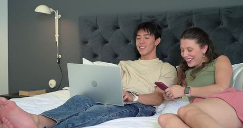 Young diverse couple relaxing on bed using laptop and smartphone in modern cozy bedroom