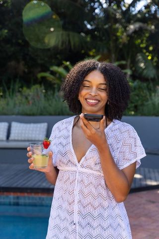 Smiling african american woman speaking into phone holding strawberry drink by sunny backyard pool