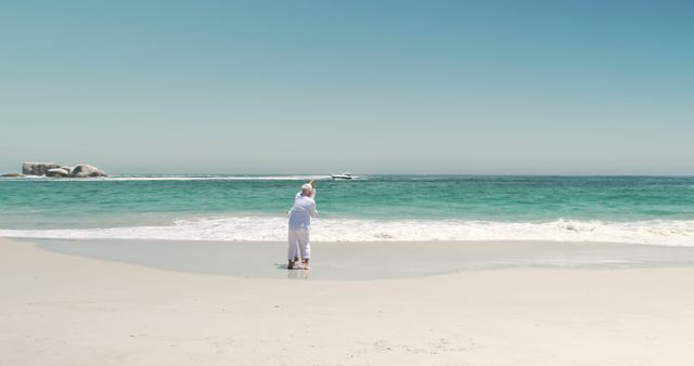 A senior couple in white clothing standing on a beach, embracing while facing the ocean. The clear blue sky and calm waves create a feeling of tranquility and peace. Suitable for use in advertising for retirement plans, travel agencies, wellness retreats, or lifestyle blogs highlighting love, relaxation, and enjoying life’s moments.
