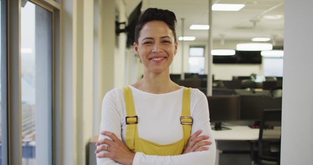 The woman in this image is in a modern office environment. She stands confidently with a smile and arms crossed, suggesting professionalism and success. This image is ideal for business, corporate, or motivational content promoting confidence, leadership, and workplace diversity.