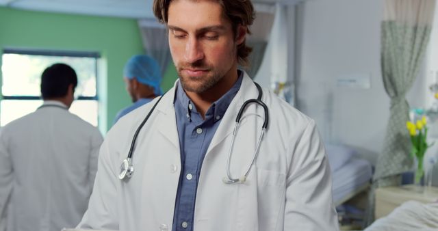 A young male doctor stands in a hospital room wearing a lab coat and stethoscope around his neck, focusing on a medical task. In the background, other medical staff and a patient bed are visible, indicating a busy healthcare setting. This image is ideal for use in healthcare, medical practice, and hospital promotional materials, illustrating professionalism and modern medical care.