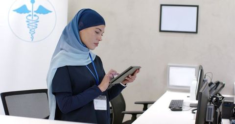 Muslim woman healthcare receptionist using tablet while standing at modern clinic desk