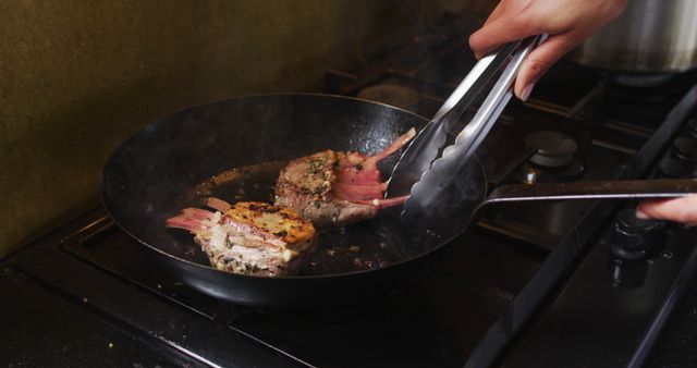 A chef is frying lamb chops in a skillet using kitchen tongs. Seasoning is evident on the meat, and steam is rising from the hot pan on the stove. This photo can be used for culinary blogs, restaurant promotions, cooking tutorials, recipe books, and advertisements for kitchen appliances or utensils.