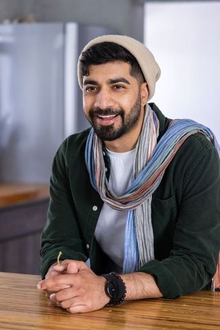 Indian man leaning on kitchen counter wearing beanie and colorful scarf, casual home portrait