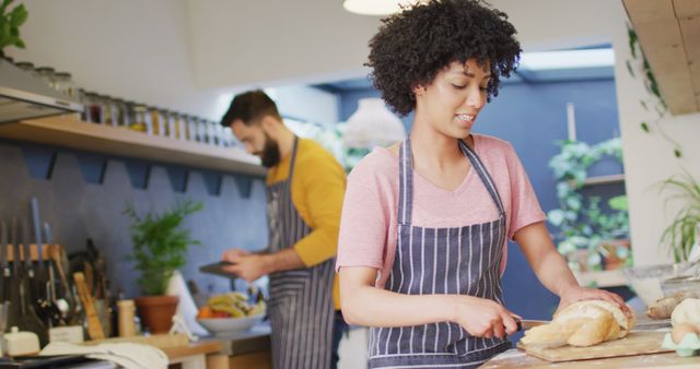 Image of happy diverse couple in aprons baking in kitchen, woman slicing bread, with copy space. Happiness, inclusivity, free time, togetherness and domestic life.