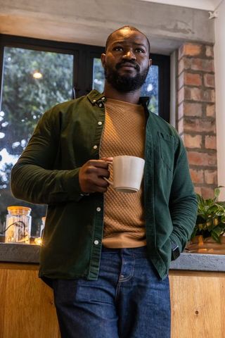 African american man holding mug and gazing out window in cozy kitchen with brick wall