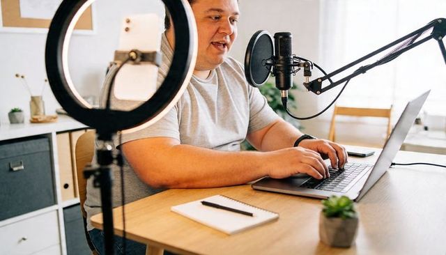 Home podcaster recording with studio microphone and ring light, typing on laptop for livestream