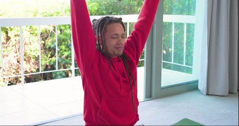 African american man stretching and meditating on yoga mat by sliding door balcony in red hoodie