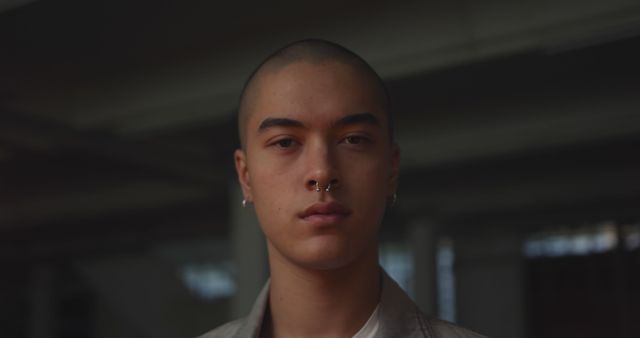 Portrait of caucasian man with shaved head, pierced nose and earrings in empty warehouse, copy space. Fashion, street style, youth, alternative and urban culture, unaltered.