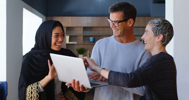 Three diverse colleagues collaborating on a project using a laptop while smiling and discussing ideas. This image can be used for marketing materials, corporate presentations, and articles about teamwork, diversity in the workplace, and professional development.