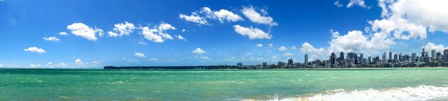 Beautiful panoramic view of Gold Coast's expansive skyline with shimmering ocean waves in the foreground under a clear, bright blue sky. Ideal for travel blogs, tourism advertisements, real estate promotions, or city guides.