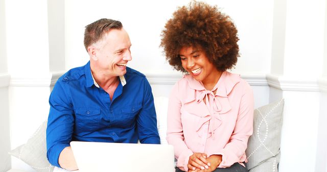 Smiling colleagues engage in a discussion over a laptop in a modern office. Perfect for illustrating concepts of teamwork, collaboration, diversity and inclusion in business settings. Ideal for use in corporate websites, training materials, promotional content, and business blogs.