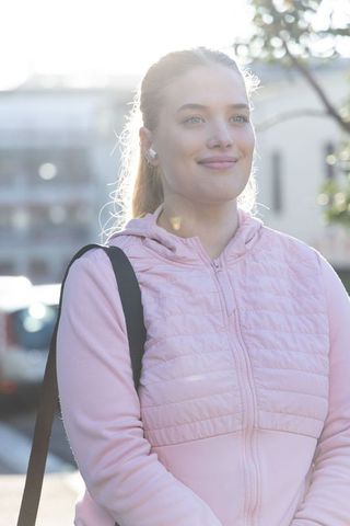 Young woman standing on urban sidewalk wearing pink quilted hoodie, earbuds, carrying bag