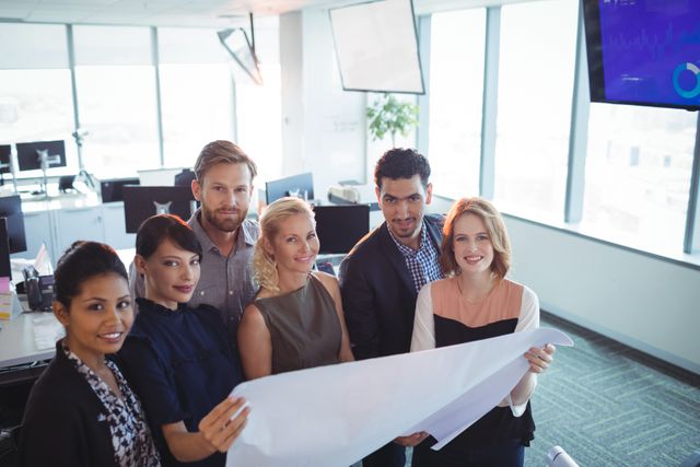 Group of diverse colleagues standing together in a modern office, holding and discussing a large blueprint. Ideal for use in business presentations, corporate websites, teamwork and collaboration concepts, and professional development materials.