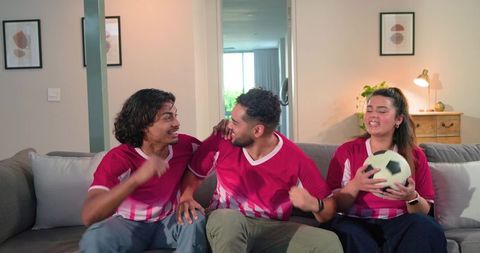 Friends cheering on couch wearing red soccer jerseys holding ball during match night