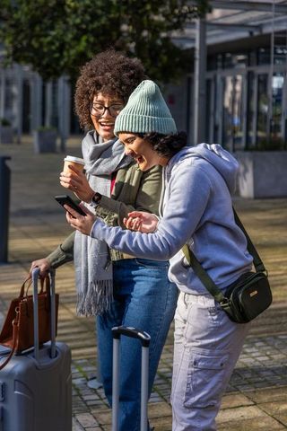 Friends laughing and checking smartphone while pulling carry-on luggage in city plaza during travel