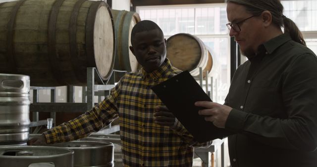 Two brewery workers are collaborating and discussing beer production in a brewery. They are next to beer barrels and kegs. One worker is holding a clipboard, and they appear to be in a deep discussion. This image can be used for articles or websites focusing on brewing industry, teamwork, manual labor, or business processes in breweries.