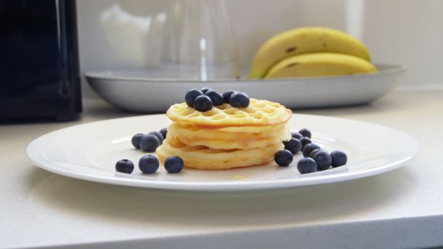 This stock video shows honey being drizzled on a stack of waffles topped with blueberries, with a plate of bananas in the background, set in a kitchen setting. The video exudes a fresh and healthy breakfast vibe. Ideal for use in culinary blogs, recipes, healthy eating promotions, kitchen-themed advertisements, and food product packaging.