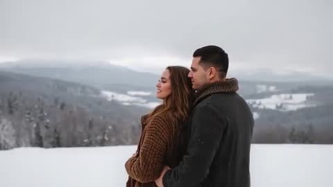Couple leaning and smiling on snowy ridge overlooking mountain valley in falling snow