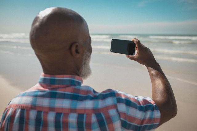 African American senior man standing on a beach, holding a smartphone and taking a photo of the sea. Ideal for use in travel blogs, vacation advertisements, technology promotions, and lifestyle articles focusing on senior activities and outdoor leisure.