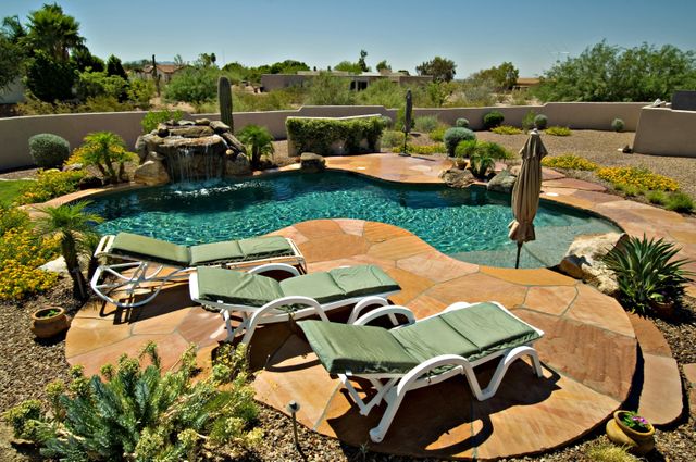 This image captures a beautifully landscaped backyard featuring a sparkling swimming pool with a waterfall, surrounded by lush greenery and desert plants. Four lounge chairs with green cushions are neatly arranged on a well-maintained patio, providing a peaceful and luxurious retreat perfect for relaxation and summer enjoyment. Ideal for advertisements for travel destinations, resorts, home improvement, and garden design inspiration.