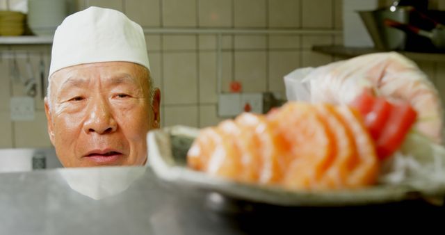 Elderly chef wearing white hat focused on preparing fresh sushi in professional kitchen. Useful for depicting culinary skills, Japanese cuisine, restaurant promotion, or articles about traditional cooking methods.