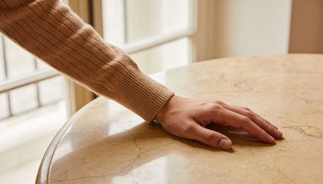 Hand resting on polished marble table wearing ribbed tan sweater by sunlit window