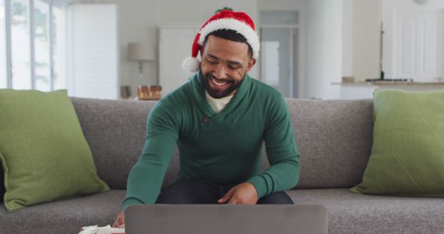 Man happily shopping online while sitting on a couch in a cozy living room setting, wearing a green sweater and Santa hat. Great for holiday season themes, online shopping promotions, festive home decor, and relaxed Christmas atmosphere concepts.