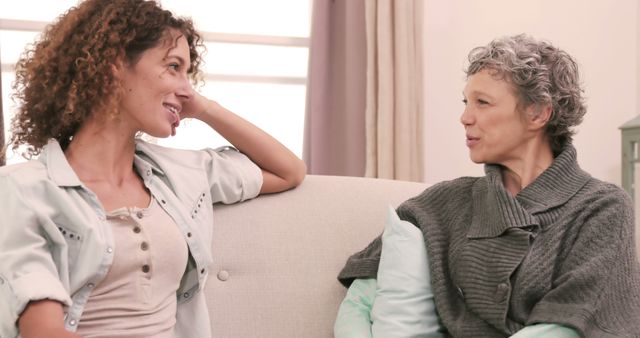 Two women are smiling and conversing on a light-colored couch in a bright and cozy room. They are engaged in a cheerful conversation, wearing casual clothing. This can be used for concepts like friendship, communication, home comfort, relaxation or casual interactions.