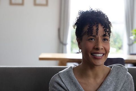 African american woman smiling and sitting on gray sofa, enjoying bright cozy living room