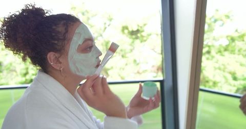 African american woman applying green facial mask on balcony wearing white bathrobe