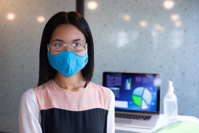 Asian woman wearing a face mask and protective eyewear standing in an office with a laptop displaying charts and graphs in the background. Useful for topics related to workplace safety, health measures during the COVID-19 pandemic, business environments, and technology use in offices.