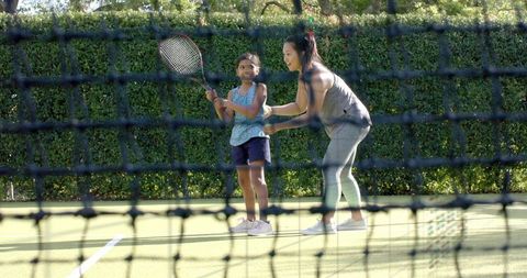 Female Coach Teaching Young Girl Tennis on Sunlit Outdoor Court with Net and Ball Basket