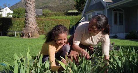 Mother teaching daughter gardening on sunny suburban lawn, tending flower bed together