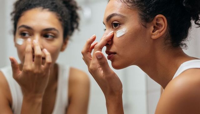 Young woman applying eye cream at mirror for morning skincare and self-care routine