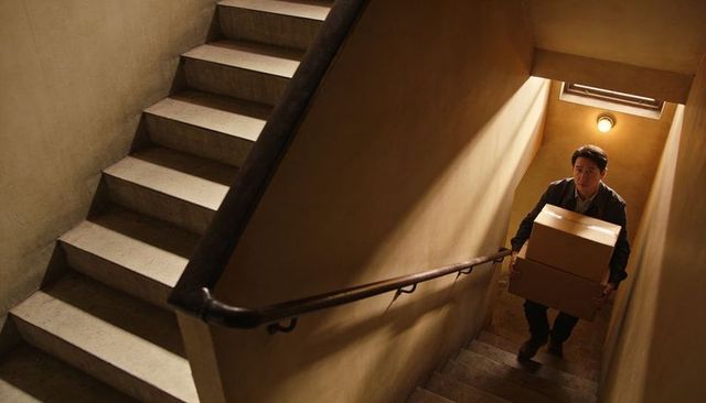 Courier carrying stacked boxes up narrow stairwell under moody warm light, delivery worker ascending