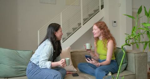Diverse women chatting and sipping mugs on sofa with smartphones in modern living space