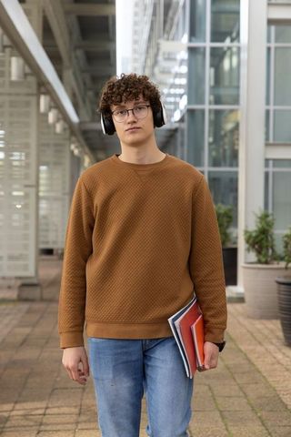 Curly-haired student standing holding notebook wearing headphones on urban campus corridor
