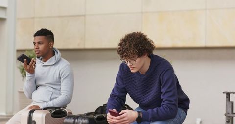 Young travelers waiting at transit terminal with suitcases and smartphones, candid urban commute