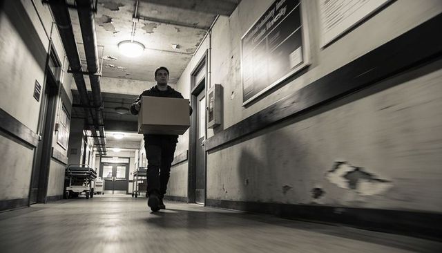 Man walking carrying box down dim hospital corridor with gurneys and peeling plaster