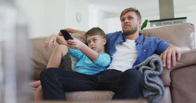 Father and son are relaxing together on a couch enjoying a TV show in the living room. This scene captures a bonding moment perfect for illustrating topics related to family life, leisure activities, home environment, and parenting. Use this for advertisements, family lifestyle articles, or home furnishing promotions.