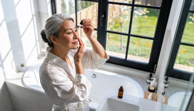 Mature woman applying facial serum at sunlit bathroom window in satin robe skincare ritual
