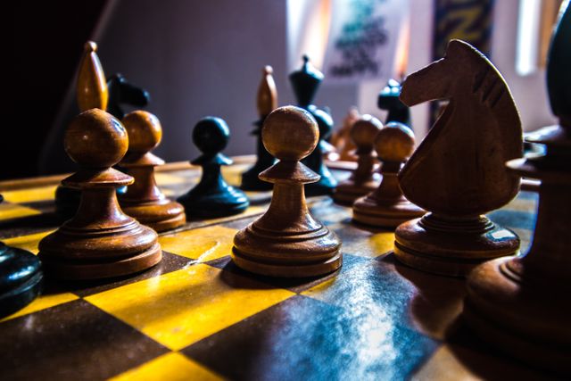 Close-up of wooden chess pieces on a chessboard, highlighted by low light entering the room. Suitable for illustrating themes of strategic thinking, competitive events, intellectual activities, or board game hobbies. Ideal for use in educational materials, strategy blogs, competition posters, or indoor activity promotions.