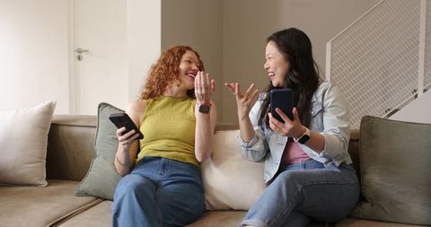 Two women laughing and chatting on sofa holding smartphones and wearing smartwatches